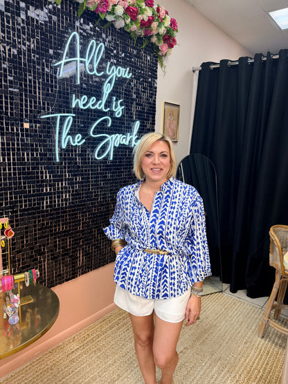 Woman in a blue patterned shirt and white shorts standing in front of a decorative wall with text.